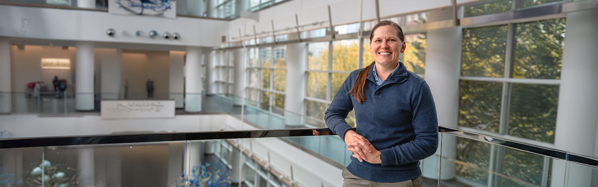 Female MBA military student, in atrium, wearing a blue sweater, smiling at camera