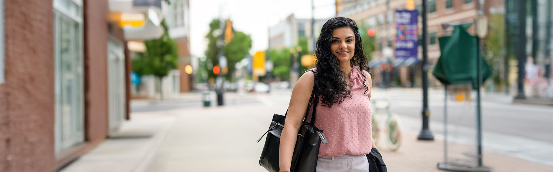 Female business student, wearing pink blouse, carrying oversized black purse, on sidewalk, looking and smiling at camera  