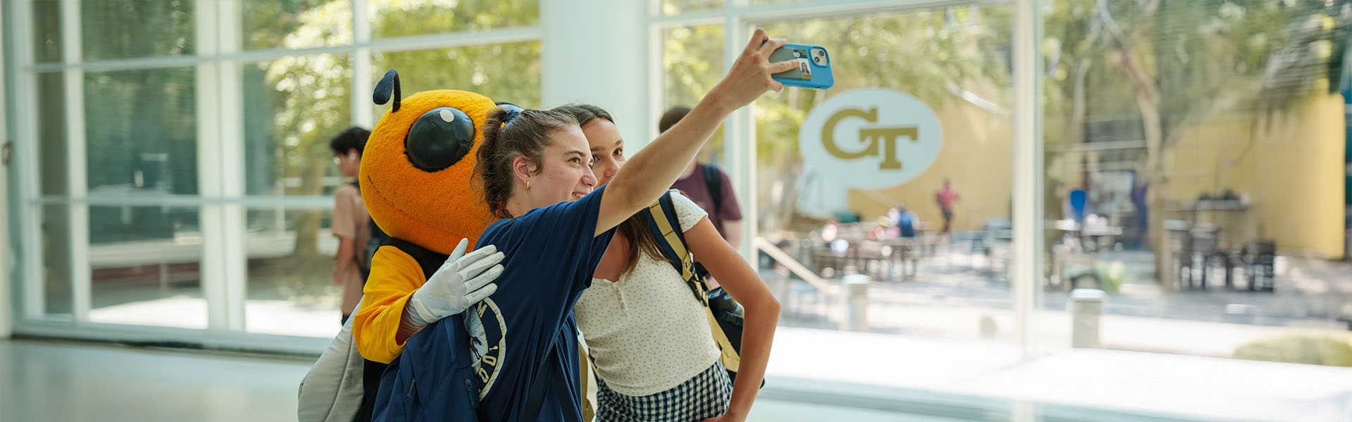 Two female undergraduate student take a selfie with Buzz in Scheller atrium