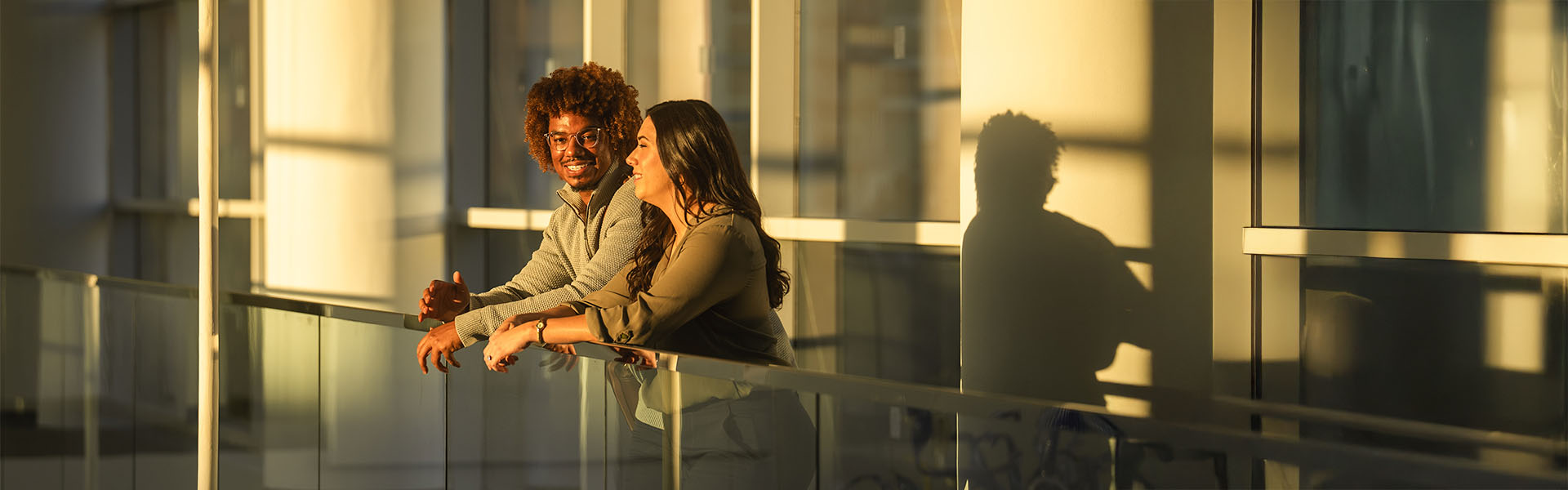 Two students leading on Scheller atrium bridge railing during sunset