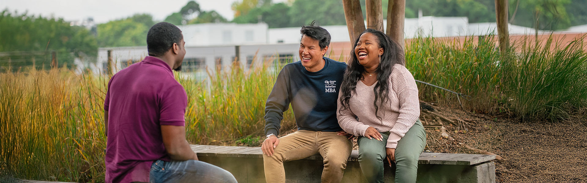 Three students laughing and interacting outside on bench in casual clothing