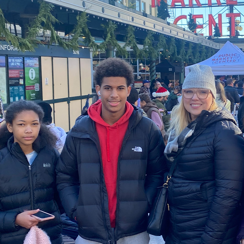 A small group of people, including two teenagers and one womn, pose in front of a Seattle, Washington farmer’s market.  