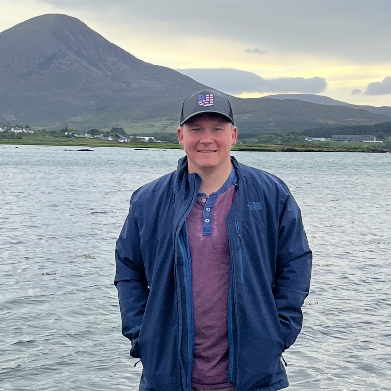 A man stands in front of a lake and mountains with his hands in his pockets