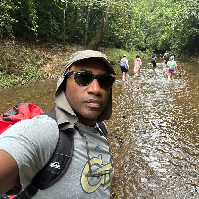 A man in a Georgia Tech t-shirt takes a selfie while walking up a creek 