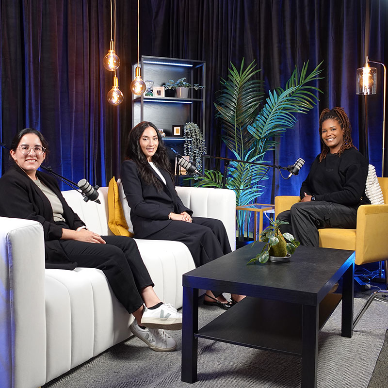 Three women sit in a room around three microphones to record a podcast.  