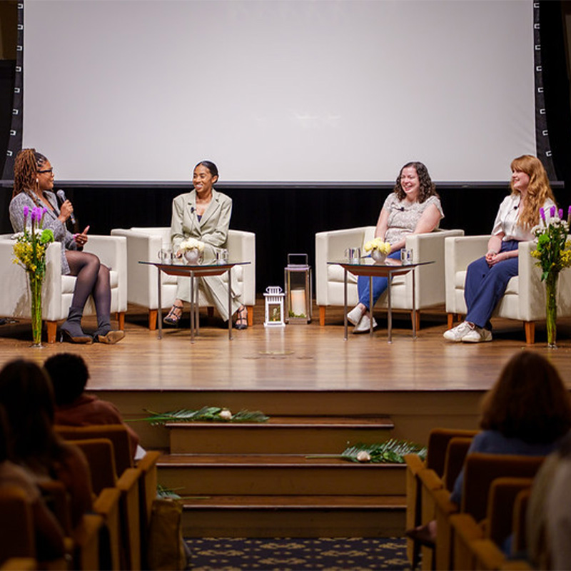 Four women sit on white chairs on a stage participating in a panel discussion.    