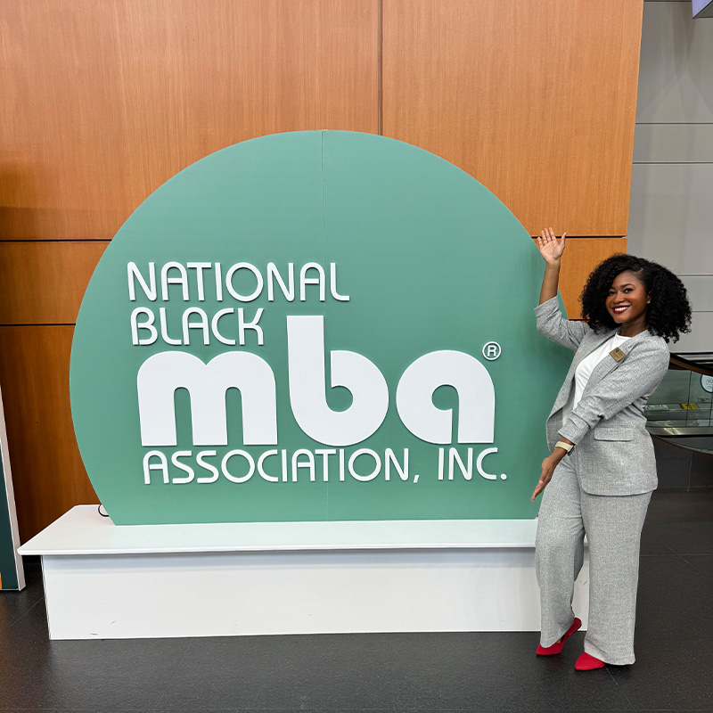 A Black female, Chasten McCrary, stands in front of the National Black MBA Association sign. 