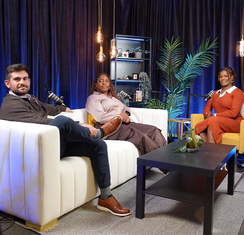 One man and two women sitting in a podcast studio with microphones. 