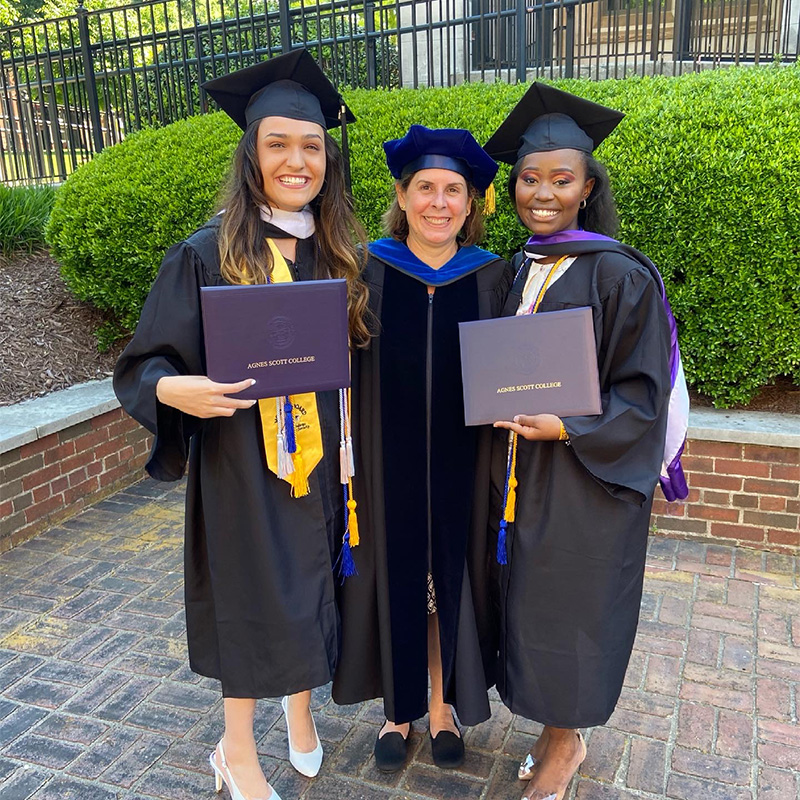 Three women in commencement regalia smile holding diplomas