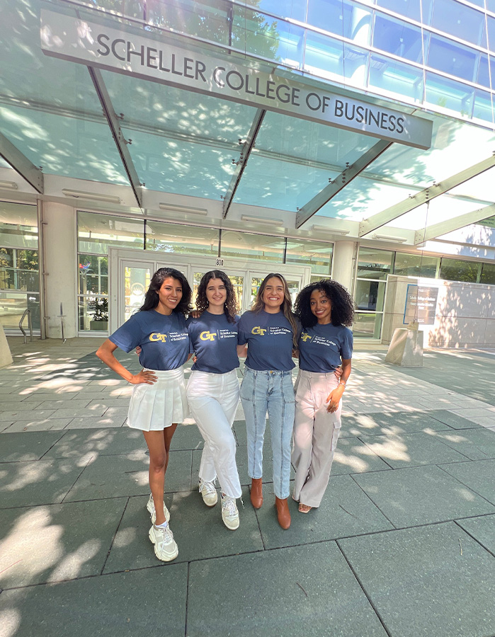 Four women in Scheller t-shirts smile in front of the college