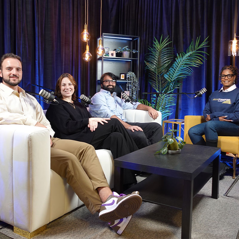 Two women and two men sit in a room with microphones to record a podcast. 