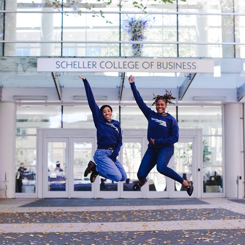 Two women in navy Georgia Tech sweatshirts jump with their right arms in the air