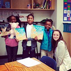 Three children holding picture books smile next to their teacher